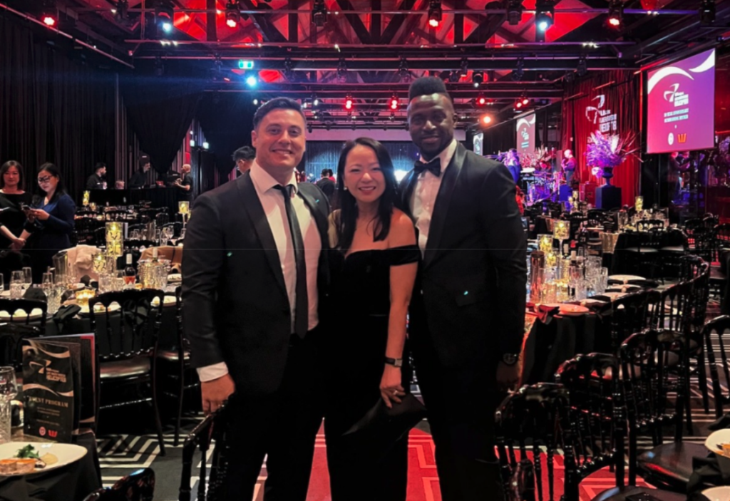 Three people dressed in formal attire stand posing for a photo at a lavish event, with decorated tables and red lighting in the background—perhaps celebrating their new business loans.